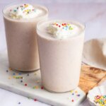 Two cake batter protein shakes in glasses topped with whipped cream and rainbow sprinkles.